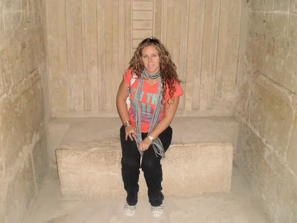 sitting in a tomb area at the Sakkara pyramids. I am dressed in a red top, black long pants and grey scarf. 