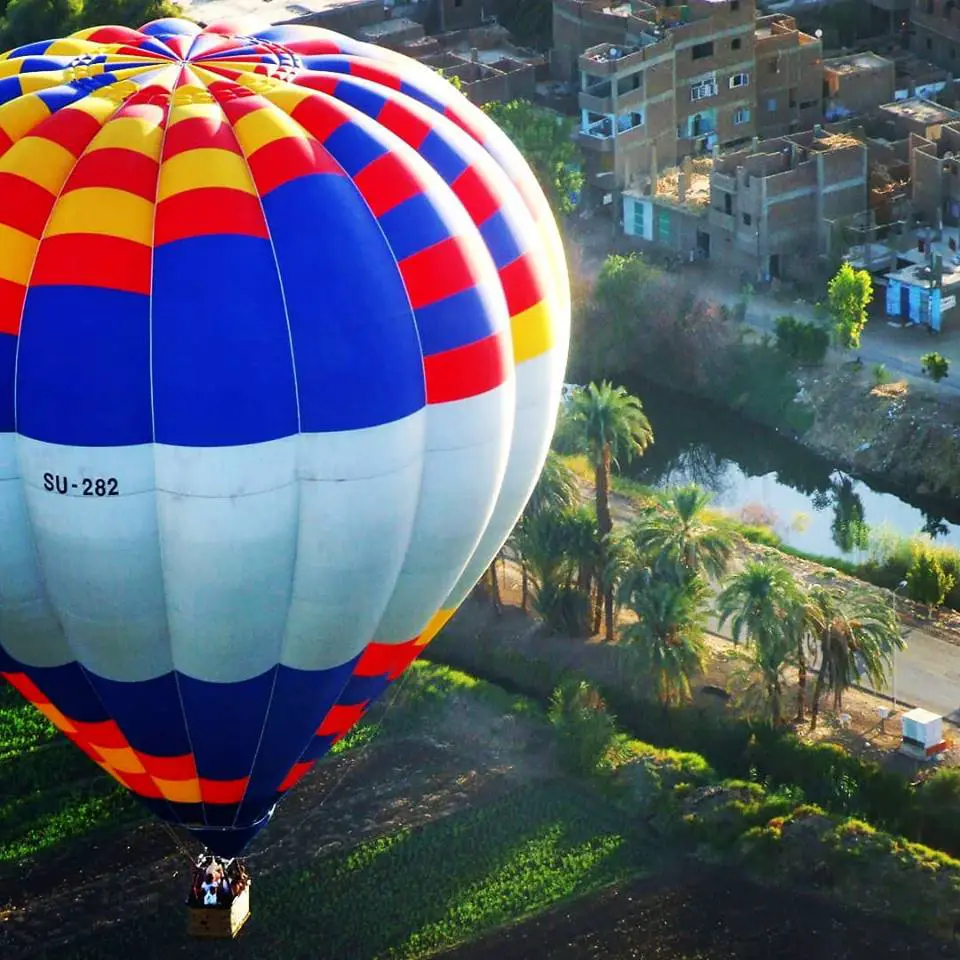 A hot air balloon with the Nile River below and house in the background. The balloon is red, white  and blue.